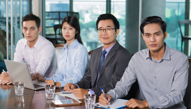 Four professionals in business attire at a meeting with laptops and notepads in an office.