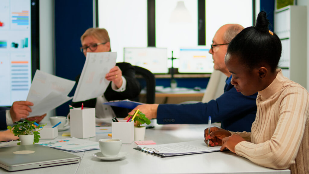 Individuals in a meeting with documents on a table, laptops, and a coffee cup visible.