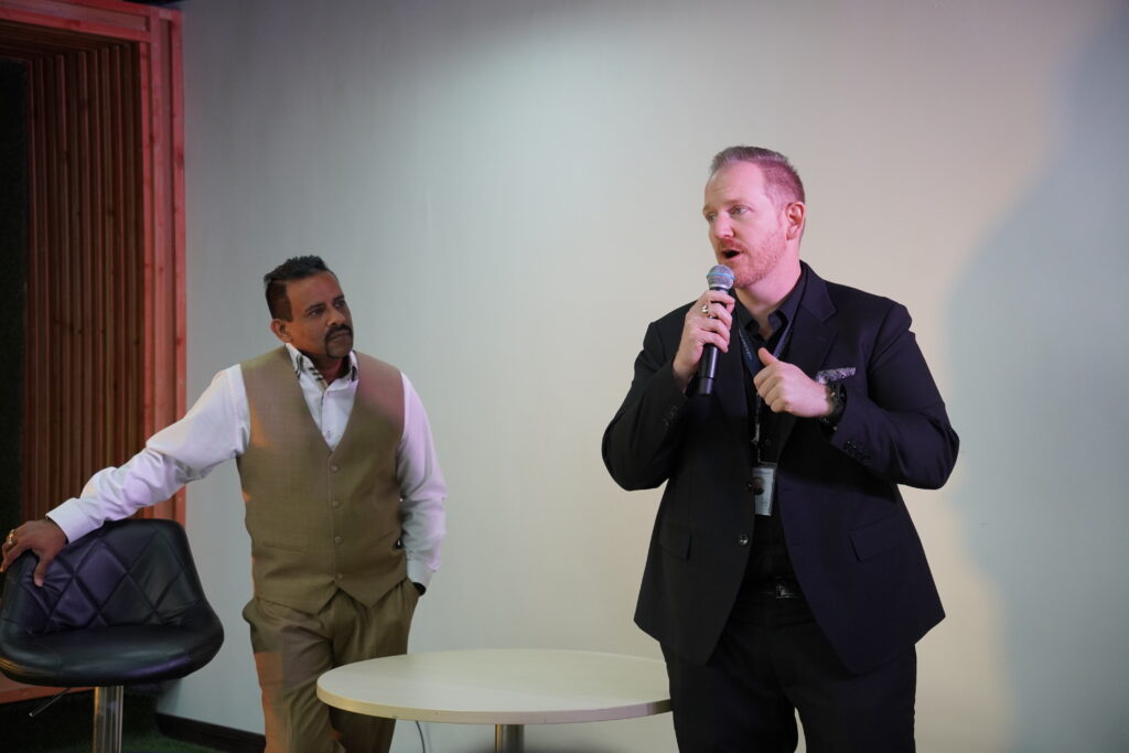 Two men in business attire, one standing by a chair and holding a microphone.