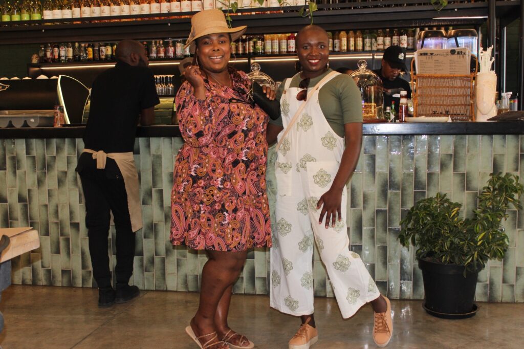 Two individuals posing in front of a cafe counter with shelves of bottles and plants.