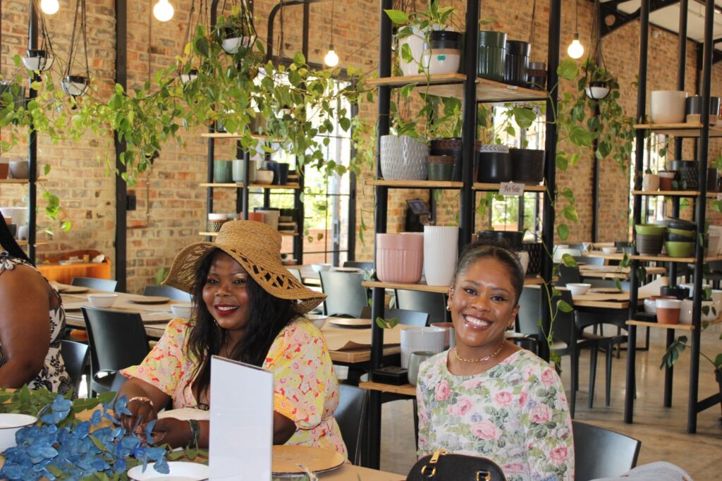 People dining in a plant-filled cafe with exposed brick walls and shelving with various pots.