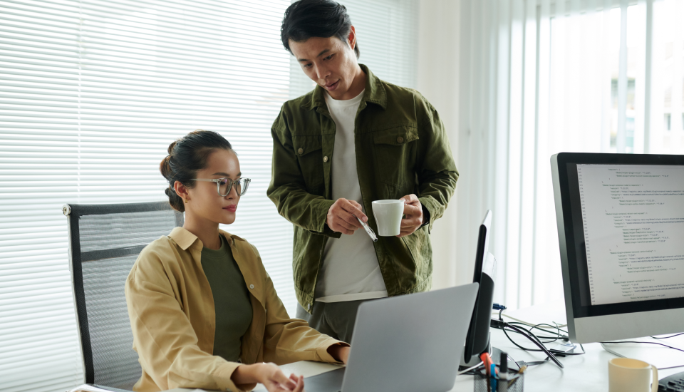 Two professionals collaborating near a laptop and desktop computer in an office setting.