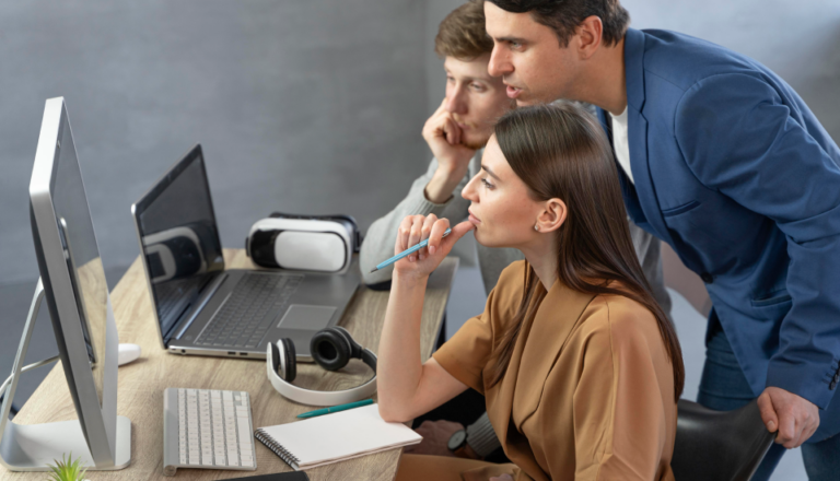 Two professionals at a desk with tech gadgets, one seated and holding a pen, the other leaning over.