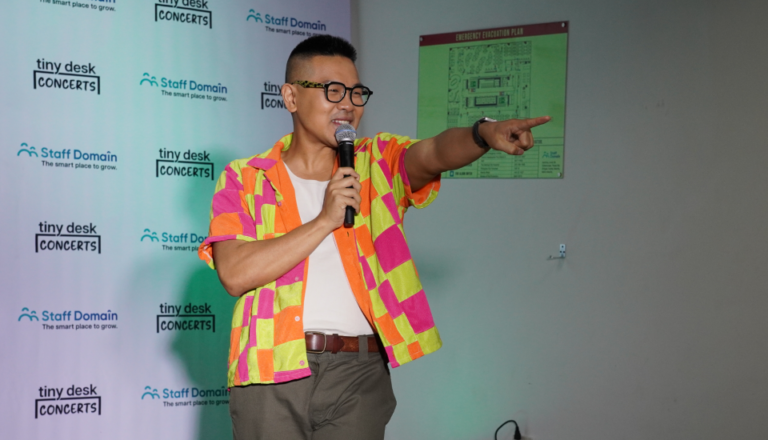 Person in a colorful shirt pointing to the side, holding a microphone, standing in front of a branded backdrop.